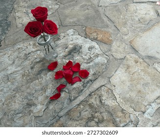Close Up Top Angle View Of Three Red Roses In A Glass Mason Jar With Silver Lid Next To Loose Pile Of Red Petals On A Grey White Stone Surface 