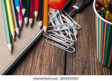A close up top down view of a collection of vibrant colored pencils and a pile of metallic paperclips arranged on a textured dark brown wooden tabletop surface