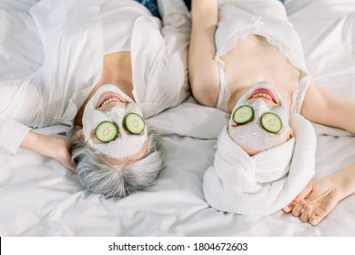 Close Up Top Image Of Women, Senior Gray Haired Lady Mother And Her Young Pretty Daughter Having Fun In The Bedroom And Making Clay Facial Masks With Cucumber Covering Their Eyes. Beauty Skin Care