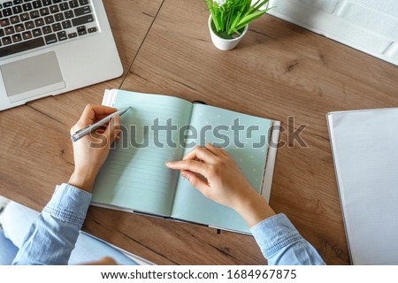 Close up top view of african american teen girl student writes notes in diary notebook journal, makes goals check list, agenda plan, organizing day concept with laptop sitting at home office desk.