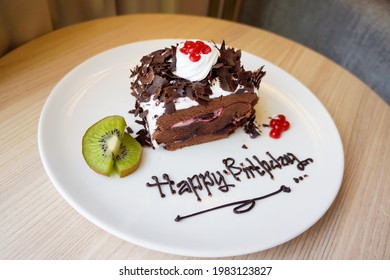 Close Up Top View Of Chocolate Birthday Cake With Whipped Cream, Sliced Kiwi And Cherry On White Plate At The Table. Bokeh Background. No People.