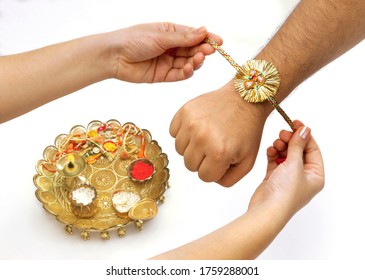 Close up top view of female  hands tying colorful rakhi on her brother’s hand with brass dish full of puja accessories in selective focus against  white background on Raksha Bandhan Festival 