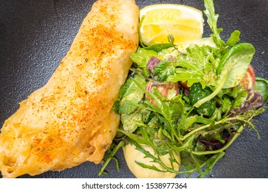 Close Up And Top View Of Fried Fish Deep Fried In Crispy Batter Served With A Side Salad Dressed, A Lemon Wedge And A Dollop Of Mayonnaise In A Black Textured Bowl For A Delicious Lunchtime Meal