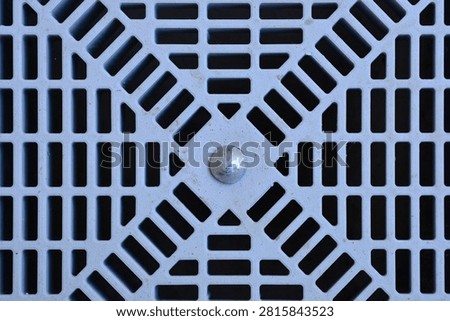 A close up top view of a grey plastic drain cover grate featuring a geometric grid pattern with rectangular slots and a central metallic screw bolt