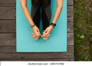 Close up Top view to hands holding foot of a female sitting on a yoga mat outdoors on a wooden floor - Powered by Shutterstock