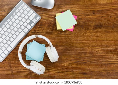 Close up top view of a keyboard, a computer mouse, headphones and small colorful sheets of paper arranged on a textured wooden surface