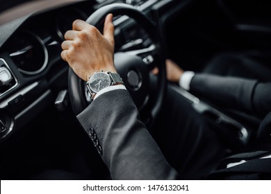 Close up top view of  man's watch in black suit keeping hand on the steering wheel while driving a luxury car.