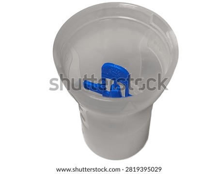 A close up top view of an open transparent plastic nebulizer medicine cup containing a blue baffle piece, with its cap placed beside it, isolated against a white background.