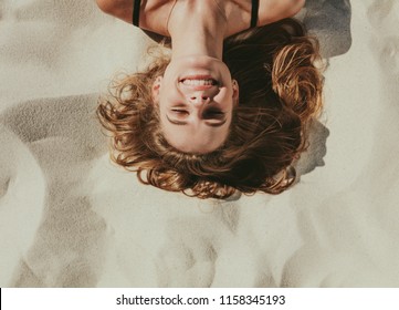 Close Up Top View Of A Smiling Woman Lying On Beach Sand. Close Up Of Head And Shoulders Of A Woman With Closed Eyes Relaxing On Beach.