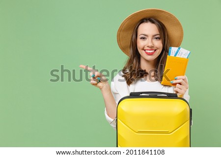 Close up tourist woman in casual clothes hat hold passport tickets yellow suitcase point finger aside workspace isolated on green background Passenger travel abroad weekend getaway Air flight concept.