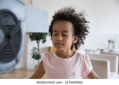 Close up tranquil little African girl stand in front of blowing fan closes eyes enjoy fresh air in living room, reduce heat inside use electric ventilator. Summer day, air-conditioner needed concept - Powered by Shutterstock