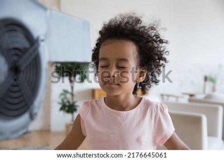 Close up tranquil little African girl stand in front of blowing fan closes eyes enjoy fresh air in living room, reduce heat inside use electric ventilator. Summer day, air-conditioner needed concept