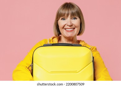 Close Up Traveler Tourist Happy Fun Mature Elderly Senior Lady Woman 55 Years Old Wears Yellow Raincoat Hold Carry Suitcase Bag In Hands Isolated On Plain Pastel Light Pink Background Studio Portrait