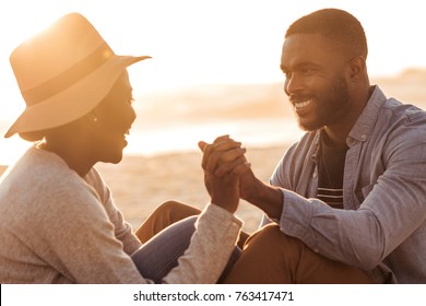 Close up of trendy young African couple sitting and holding hands together on a sandy beach while enjoying a romantic sunset - Powered by Shutterstock