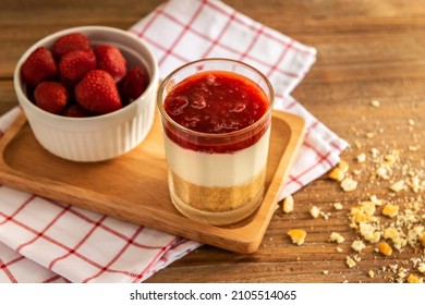 Close up of triple crispy strawberry cheesecake in shot glass on square wooden tray over red, white plaid napkin with some crackers crumble on the table. Eatable recipe decoration dessert concept.