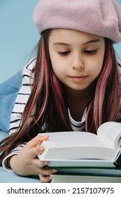 Close Up Of A Tween Girl In French Beret Reading Books, Lying On The Floor. She Has Dyed Hair Strands. Over Blue Background.