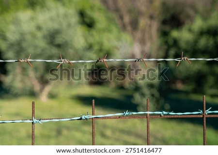 Close up of twisted barbed wire stretched across a fence with rusty metal posts and a second wire below, sharp focus on barbs and knots with shallow depth of field and soft green countryside blur behind
