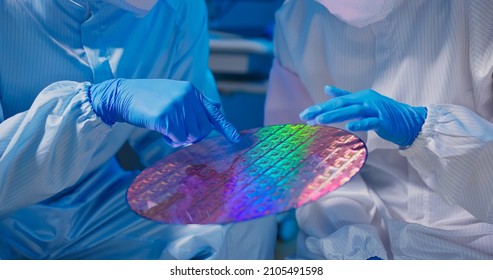 close up of two asian technicians in sterile coverall hold wafer with gloves that reflects many different colors and check it at semiconductor manufacturing plant