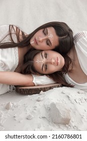 Close Up Two Brunette Girls In White Boho Style Dresses Are Lying Head On Head With Closed Eyes And Make Up On A Sand Career Background In Desert. Modern Shamans. Lifestyle Fashion Concept, Free Space