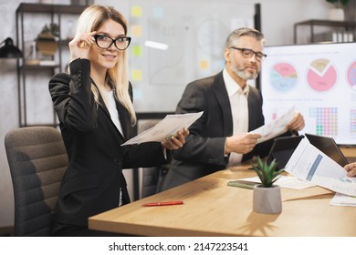 Close up of two business colleagues at the table in modern office room, working together with digital tablet and papers. Focus on young blond woman, smiling at camera while making notes. - Powered by Shutterstock