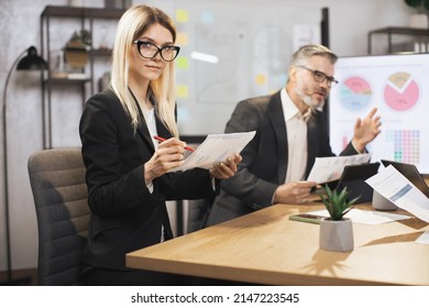 Close up of two business colleagues at the table in modern office room, working together with digital tablet and papers. Focus on young blond woman, smiling at camera while making notes. - Powered by Shutterstock