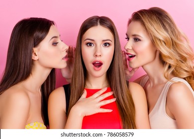 Close up of two charming girls holding hand near mouth telling gossips on ears to their shocked friend with open mouth who giving impressed, unexpected reaction while standing over pink background