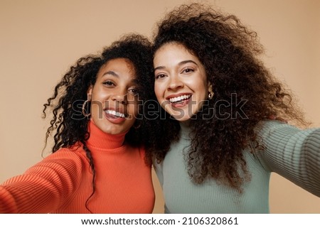 Close up two cheerful happy beautiful young curly black women friends 20s wearing casual shirts clothes doing selfie shot pov on mobile phone isolated on plain pastel beige background studio portrait