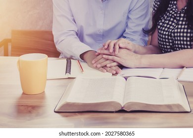 Close Up Of Two Christian Women Studying The Bible, Holding Hands, And Pray Together Around A Wooden Table In The Home, Christian Discipleship Concept. Devotional Background With Copy Space.