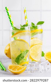 Close up of two clear glasses with refreshing homemade lemonade with fresh lemon slices, mint leaves, ice cubes and paper straws on a light kitchen counter against white tile background