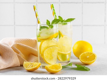 Close up of two clear glasses with refreshing homemade lemonade with fresh lemon slices, mint leaves, ice cubes and paper straws on a light kitchen counter against white tile background