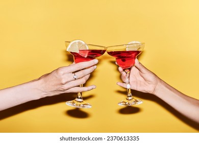 Close up of two female hands holding red cocktails against vibrant yellow background, copy space