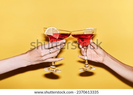 Close up of two female hands holding red cocktails against vibrant yellow background, copy space