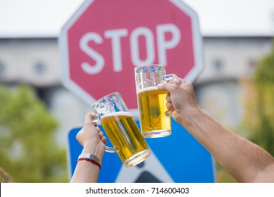 Close Up Of Two Hands Holding Mugs Of Beer In Front Of Stop Street Sign. Alcohol Abuse And Alcoholism Prevention. Don't Drink And Drive Concept