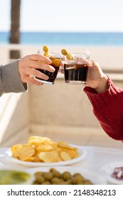 Close up of two hands toasting with their glasses of vermouth on a table full of snacks in front of the beach during a sunny day. Two people with no faces, toasting with some drinks with copy space.