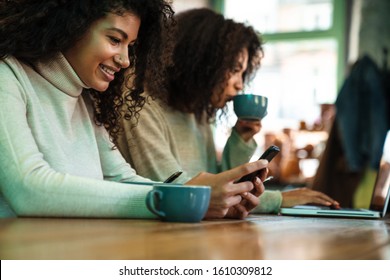 Close up of two happy african girlfriends sitting at the cafe, having cup of tea, working on a laptop, using mobile phone