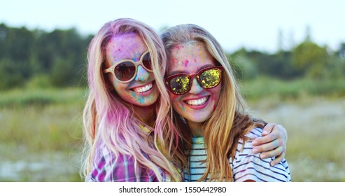 Close up of the two joyful and beautiful Caucasian blond yung women in glasses looking at each other and smiling to the camera, best friends having fun at the holi fest. Portrait. Outdoor.