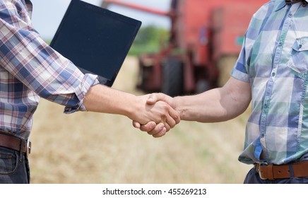 Close up of two men in plaid shirts shaking hands on the farmland. Combine harvester working in background