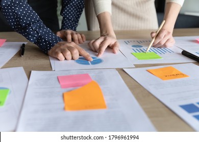 Close up two multiracial business women analyzing marketing research charts on paper document, developing economic growth strategy, managing working processes using memo colorful stickers in office.