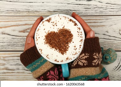 Close up two woman hands hold and hug big full cup of latte cappuccino coffee with heart shaped chocolate on milk froth over white wooden table, elevated top view, directly above