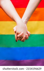 Close Up Of Two Woman's Hands Holding Each Other With The LGBTQ Rainbow Flag In The Background. Symbol For Homosexual Community For Pride Month And The Celebration Day Of Sexual And Love Diversity