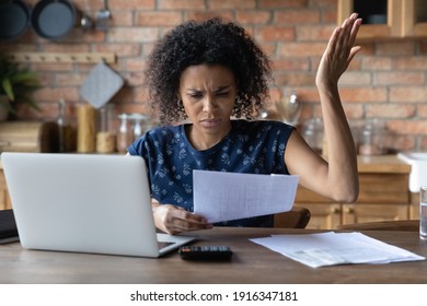 Close up unhappy African American woman calculating bills, irritated by financial problem, unexpected debt, bankruptcy, holding document or receipt, lack of money, sitting at table with laptop