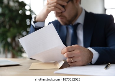 Close up unhappy frustrated businessman holding paper letter, sitting at work desk in office, upset stressed young employee executive wearing glasses received bad news, dismissal, unpaid debt