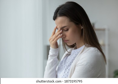 Close up unhappy stressed young woman standing near window at home, touching nose bridge, suffering from headache or migraine, thoughtful melancholic girl thinking about problems, lost in thoughts - Shutterstock ID 1873195795