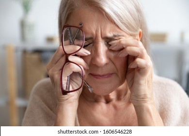 Close up of unhealthy elderly woman take off glasses massage eyes suffering from strong migraine or headache, unwell sick senior female grandmother struggle with blurry vision or dizziness at home