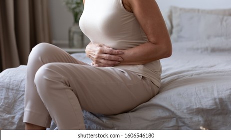 Close up of unhealthy senior woman sit on bed at home touch hold belly suffer from stomach ache, sick mature female struggle with diarrhea or food poisoning, having gastritis or abdominal pain