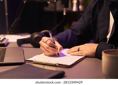 Close up of unknown man writing notes on clipboard beside laptop and headphones, managing workflow at desk. Suitable for productivity, remote work, planning or content creation marketing