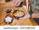 Close up of unrecognizable chef adding seeds with a small spoon to healthy breakfast bowl made with mango, banana, raspberries, cereals and muesli during a cooking master class. Vegan meal concept.