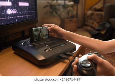 Close up of unrecognizable child inserting cartridge into vintage console while holding controller in hand with old-fashioned game visible on TV screen