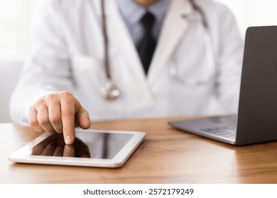 Close up of unrecognizable MD using tablet and computer, touching screen, sitting at wooden desk