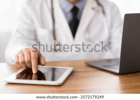 Close up of unrecognizable MD using tablet and computer, touching screen, sitting at wooden desk